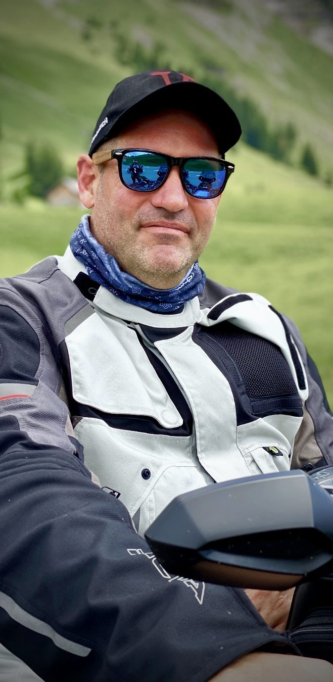 Tom´s Portrait Mann in Motorradjacke und Sonnenbrille, sitzt auf seinem Motorrad inmitten einer grünen Berglandschaft.